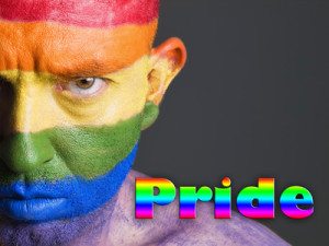 Gay flag painted on the face of a man. Man is looking at camera and has a serious expression. The word Pride is written at one side.