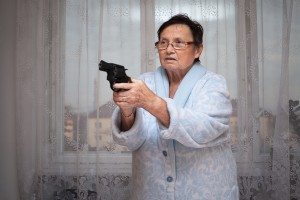 Scared senior woman aiming a gun indoors.