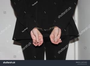 Person in a black suit with hands cuffed behind their back in front of a plain wall, facing the camera.